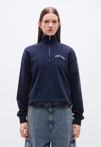 Μπλε ναυτικό cropped φούτερ με ψηλό γιακά, μισή φερμουάρ και λευκό λογότυπο "TOMMY JEANS" στο στήθος. Μαλακή υφή.