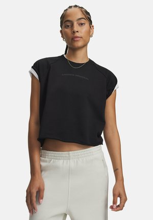 Vrouw met gevlochten haar draagt een zwart, kort mouwloos topje en lichtgekleurde broek met elastische taille, staand tegen een effen achtergrond.