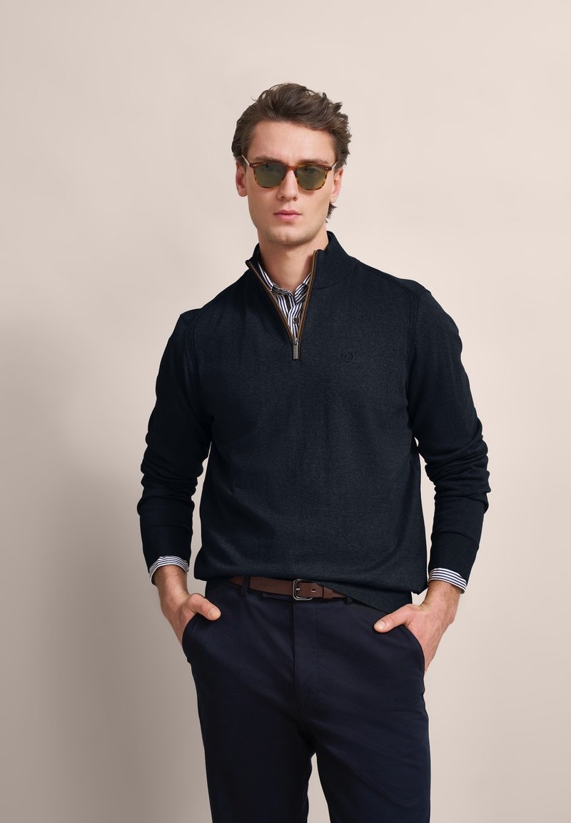 Dark navy sweater with a quarter-zip design, ribbed cuffs, and a patterned collar underneath. Model wears green-tinted sunglasses.