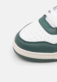 Zbliżenie na biały i ciemnozielony czubek sneakersów z perforowanym białym skórzanym materiałem i białymi sznurowadłami na jasnym tle.