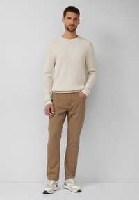 Mann trägt einen cremefarbenen Strickpullover, hellbraune Hose und weiße Turnschuhe und steht mit einer Hand in der Tasche vor einem hellgrauen Hintergrund.