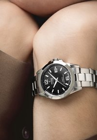 Orologio in acciaio inox argentato con quadrante nero, dotato di indici ore bianchi, display della data e bracciale a maglie metalliche.