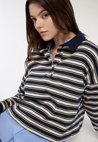 Polo a maniche lunghe a righe blu navy e crema, con colletto scuro a contrasto, dotato di patta con bottoni e morbido tessuto di cotone.