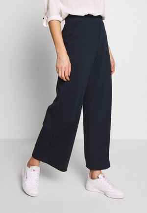 Personne portant un pantalon noir taille haute à jambes larges, associé à des baskets blanches et une chemise blanche à manches longues avec les manches retroussées.