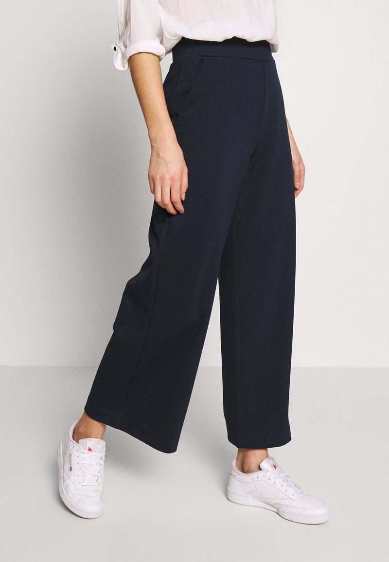 Personne portant un pantalon noir taille haute à jambes larges, associé à des baskets blanches et une chemise blanche à manches longues avec les manches retroussées.