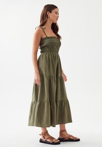 Frau trägt ein olivgrünes, gestuftes Midikleid mit gepufftem Oberteil und Spaghettiträgern, kombiniert mit braunen flachen Sandalen.