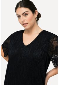 Mujer con el pelo oscuro recogido en un moño, viste una blusa de encaje negro con mangas cortas y pendientes de aro, mirando ligeramente hacia un lado con una expresión serena.