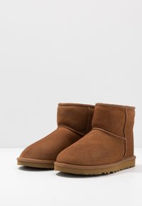 Botine din suede maro la glezna, cu vârful rotunjit, textură moale, cusături decorative și talpă din cauciuc cu un model de profil.