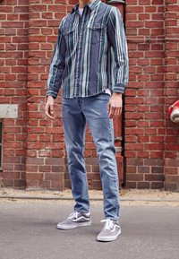 Camisa de botones a rayas en verde y azul marino, combinada con jeans de color azul claro y zapatillas grises. Colocada frente a un fondo de pared de ladrillo.
