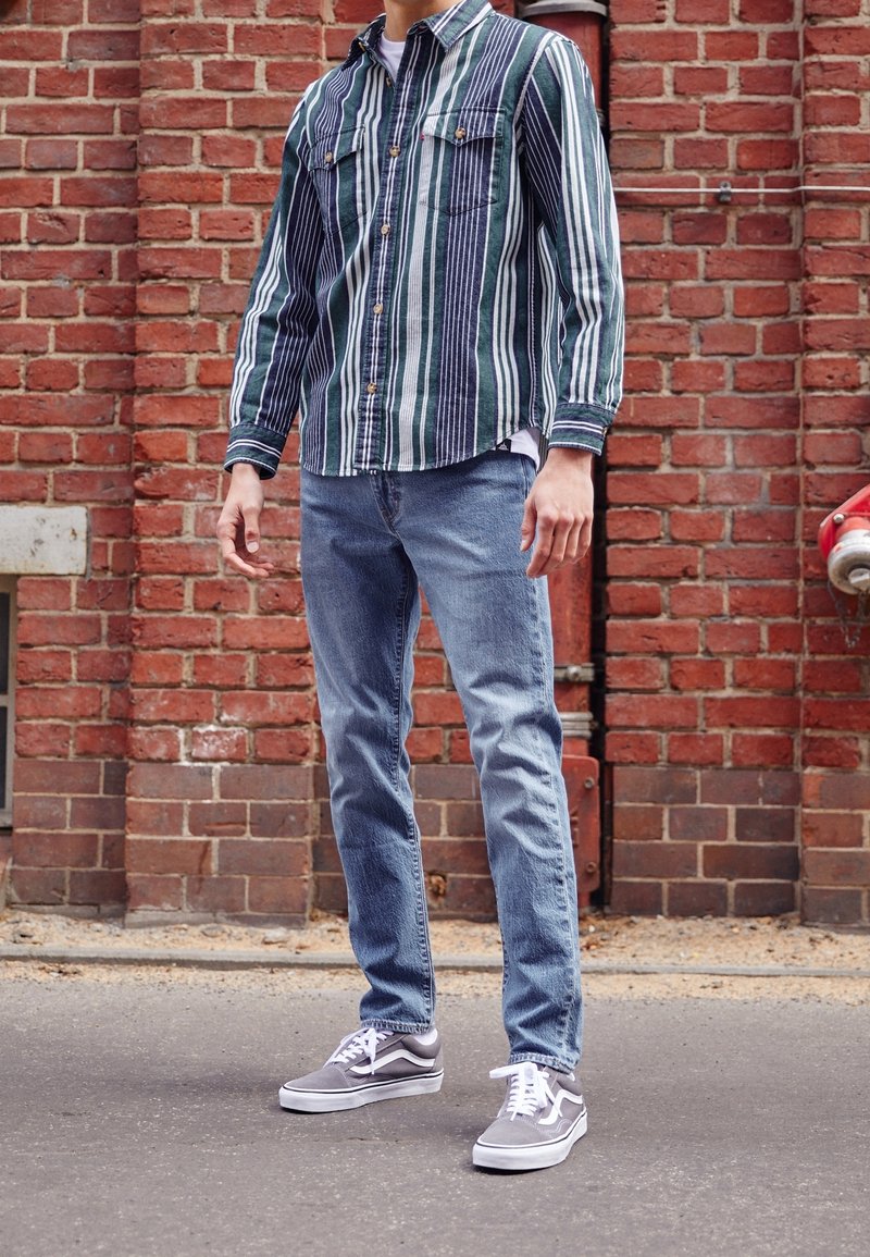 Camisa de botones a rayas en verde y azul marino, combinada con jeans de color azul claro y zapatillas grises. Colocada frente a un fondo de pared de ladrillo.