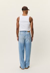 Λευκό αμάνικο τοπ, ανοιχτό μπλε τζιν με άνετη εφαρμογή, ορατές ραφές και δερμάτινη ετικέτα στη πίσω μέση. Καφέ καπέλο φορεμένο ανάποδα.