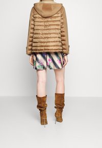 Pufferjakke i tan med en blød hætte, udstyret med et quiltet design. Sammen med et blomster-mønstret nederdel og brune slouchy knæ-høje støvler.