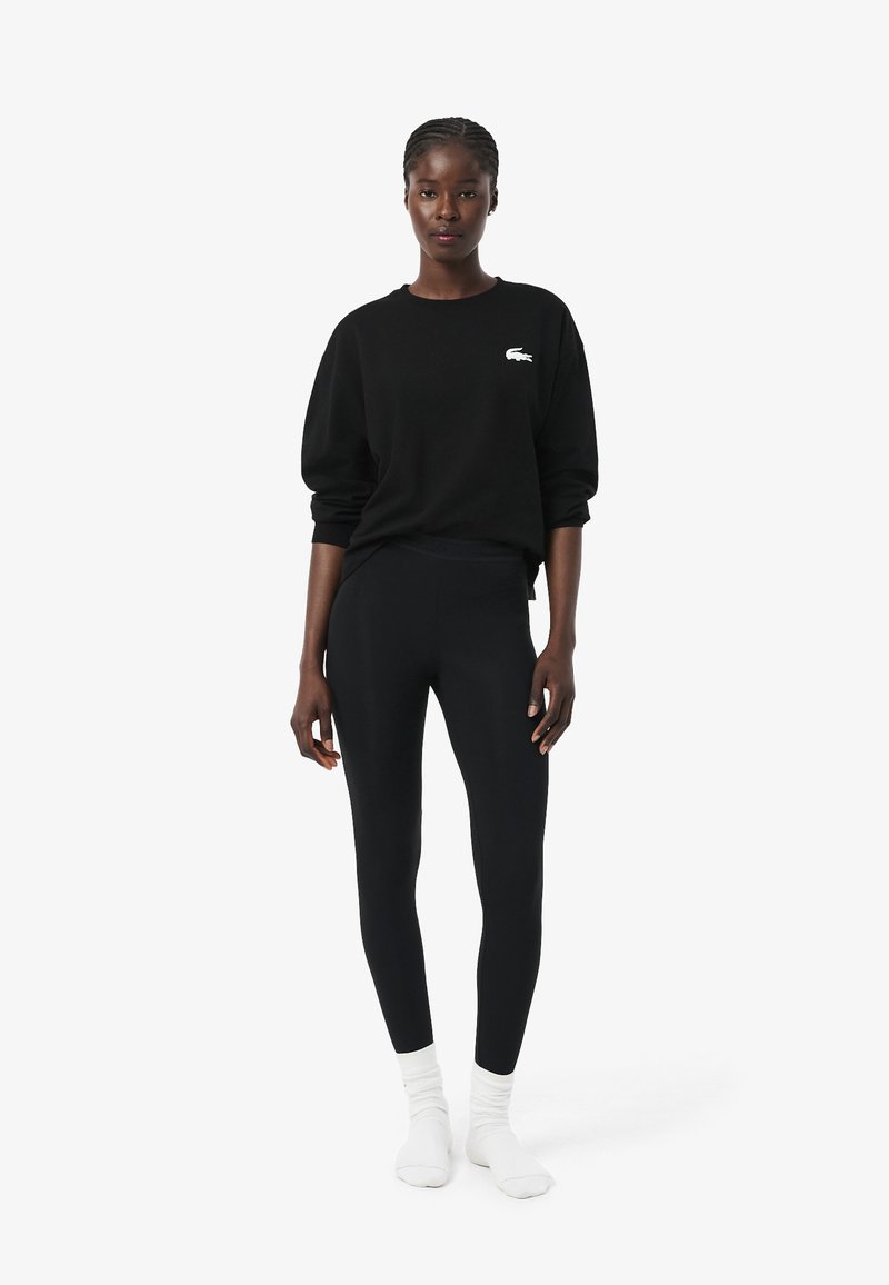 Vrouw draagt een zwarte trui met een wit logo, zwarte legging en witte sokken, staand tegen een effen witte achtergrond.