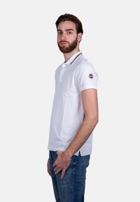 Polo shirt bianco a maniche corte con colletto, caratterizzato da una striscia a contrasto, patch con logo sulla manica sinistra e una vestibilità dritta. Jeans in denim indossati.