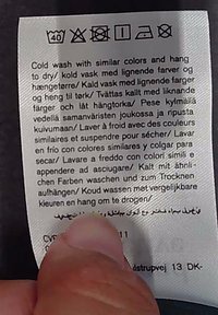 Waschetikett mit Waschsymbolen und mehrsprachigen Pflegeanweisungen zum Kaltwaschen mit ähnlichen Farben und zum Aufhängen zum Trocknen, gehalten von einem Daumen.