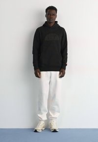 Sudadera con capucha negra y diseño en relieve, combinada con pantalones de chándal blancos. Modelo lleva zapatillas blancas con acentos oscuros. Fondo liso.