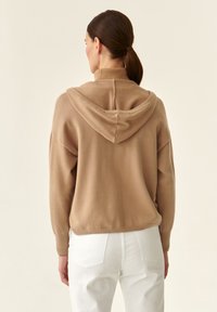 Maglione beige con cappuccio in morbido tessuto a maglia, dotato di un colletto alto, spalle scese e polsini a coste, abbinato a pantaloni bianchi.