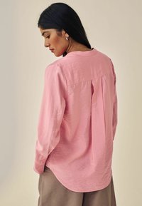 Maglietta rosa a maniche lunghe con finitura texture, scollo tondo e dettagli di pieghe sul retro. Abbinata a pantaloni in colori neutri.