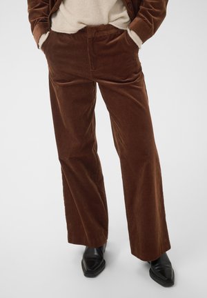 Personne portant un pantalon large en velours côtelé marron, un pull beige et des bottes noires à bout pointu, les mains dans les poches.