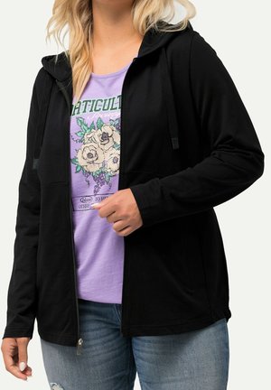 Kobieta ubrana w czarną bluzę z kapturem i zamkiem błyskawicznym na lawendowym t-shircie z kwiatami i napisem, w połączeniu z jasnoniebieskimi jeansami.
