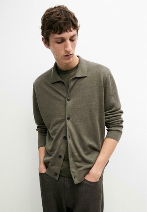 Giovane uomo che indossa un cardigan verde oliva con bottoni sopra una camicia abbinata, con le mani nelle tasche dei pantaloni scuri, guarda verso il basso, su sfondo bianco uniforme.