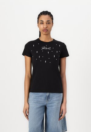 T-shirt noir à manches courtes avec un design ajusté, arborant des embellissements argentés et un logo au centre, associé à un jean bleu clair.