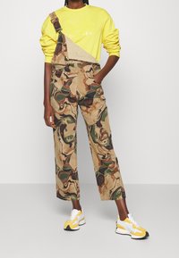 Sudadera amarilla con un gráfico impreso, combinada con un mono de camuflaje de piernas amplias. Unas zapatillas blancas y amarillas completan el conjunto.