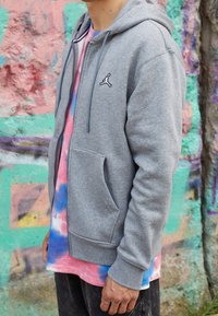 Felpa grigia con zip e coulisse, con il logo Jordan nero. Indossata sopra una maglietta tie-dye rosa e blu, esposta su uno sfondo colorato.