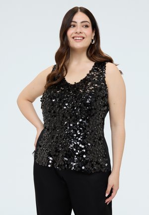 Mujer con el cabello largo y castaño, vistiendo una camiseta sin mangas negra cubierta de grandes lentejuelas brillantes, sonriendo y posando con una mano en la cadera.