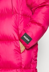 Chaqueta acolchada color rosa brillante con diseño acolchado. Cuenta con una etiqueta negra con la palabra "HUGO" y una capucha con cordón ajustable. Textura suave.