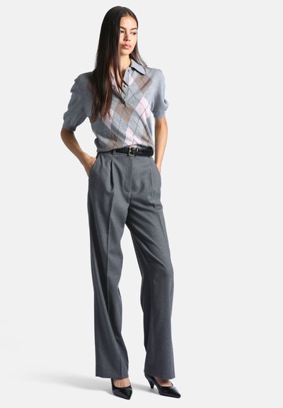 Femme debout les mains dans les poches, portant un pull gris à manches courtes avec motif losange, pantalon gris taille haute, ceinture noire et talons noirs.