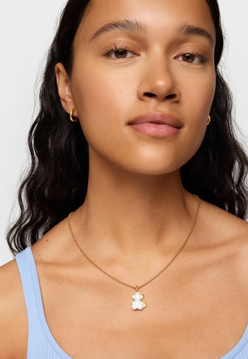 Collier en chaîne dorée avec un pendentif en forme d'ours de couleur blanche. L'ours présente un accent doré, porté sur un haut bleu clair. Texture lisse.