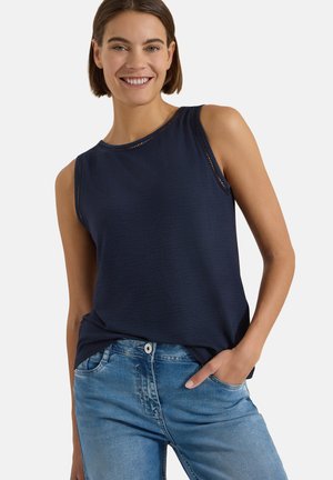 Jeune femme souriante portant un haut sans manches bleu marine avec garniture décorative et un jean bleu clair, une main dans la poche, sur fond uni.