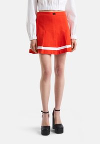 Röd plisserad kjol med vita ränder vid fållen, med resårmidja och en svart emblem. Kompletterad med svarta platåskor.