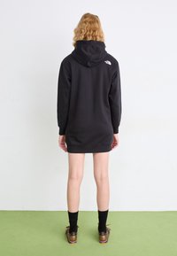 Personne aux cheveux blonds bouclés, vue de dos, portant une robe à capuche noire avec un logo blanc dans le haut du dos, des chaussettes noires et des chaussures marron.