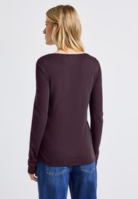 Långärmad topp i djupt lila tyg med rund halsringning, som har en slät textur och en lätt kurva vid nederkanten. Bärs med blå jeans.