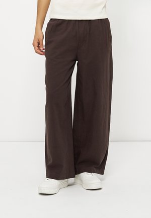Personne portant un pantalon large ample couleur brun foncé, des baskets blanches et un haut blanc sans manches, debout devant un fond blanc uni.