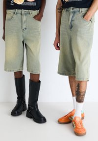 Shorts en denim vert clair avec une coupe décontractée, associés à des bottes noires montantes et des baskets en daim orange avec des accents blancs et des chaussettes.