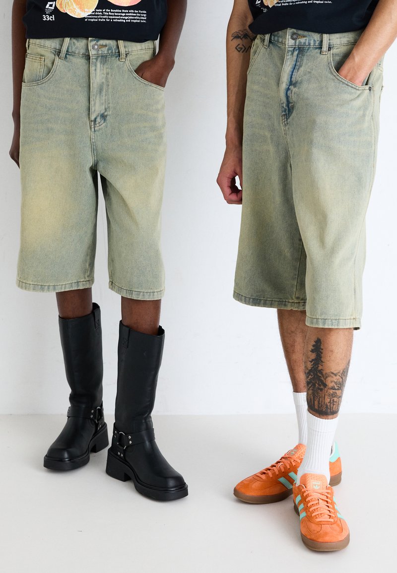 Shorts en denim vert clair avec une coupe décontractée, associés à des bottes noires montantes et des baskets en daim orange avec des accents blancs et des chaussettes.