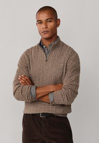 Bruine geribbelde gebreide pullover met een ritskraag, over een lichtgrijs shirt gedragen. Donkere corduroy broek en een zwarte riem zichtbaar.