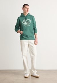 Hanorac verde cu glugă, text grafic alb, buzunar tip cangur, asortat cu pantaloni albi și adidași albi cu talpă bej, pe un fundal neutru.