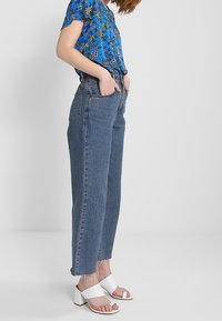 Modrá květinová košile s krátkým rukávem, světle modré džíny s vysokým pasem a bočními kapsami, a bílé sandály na podpatku se zapínáním.