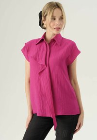 Blusa rosa a maniche corte con superficie texture, caratterizzata da una chiusura con bottoni frontalmente e dettagli arricciati lungo il colletto. Abbinata a pantaloni neri.