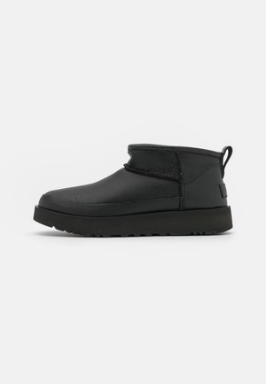 Bottine en cuir noir avec une semelle plate texturée, une languette au talon et un col rembourré, vue de profil sur un fond blanc.