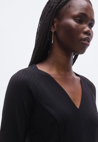 Mustast soonikangast topp V-kaelusega, millel on pikad varrukad ja tekstuuriga materjal. Kõrvarõngad ripuvad ja lisavad peent detaili.