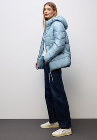 Ljusblå pufferjacka med huva, med kviltad textur och elastiska detaljer. Kombinerad med mörkblå jeans och vita sneakers.