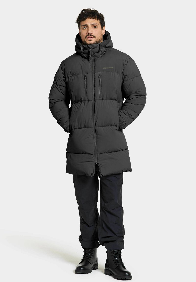 Schwarze, gepolsterte Parka mit hohem Kragen, Reißverschluss vorn und zwei Brusttaschen. Wird über schwarzen Hosen und Stiefeln getragen, die eine glänzende Textur aufweisen.