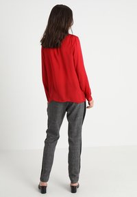 Blouse rouge à manches longues avec épaules froncées, associée à un pantalon gris à motifs avec une bande noire sur le côté. Texture lisse.