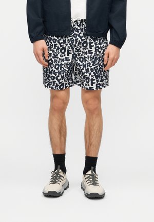 Mężczyzna ubrany w czarno-białe spodenki z wzorem liter, czarne skarpetki, białe trampki, ciemną kurtkę i białą koszulę, stojący z rozluźnionymi rękami.