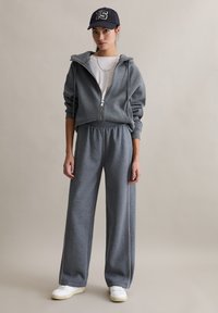 Szara bluza na zamek z krótkim krojem, zestawiona z szarymi, szerokimi spodenkami dresowymi. Oba elementy mają miękką teksturę i elastyczny pas. Białe akcenty na spodniach.
